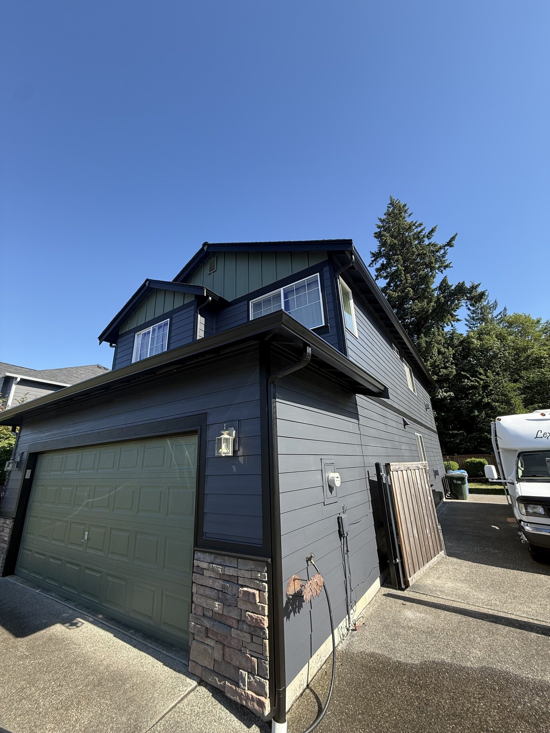 Board-and-batten home with stone accents, dark gutters, and downspout, with mature firs behind