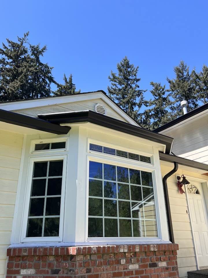 Dark seamless gutters installed on a bay window with Pacific Northwest fir trees